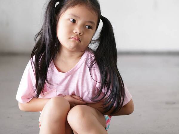 A young girl with pigtails sits on the ground with a look on her face that she doesn't feel well as she holds her stomach.
