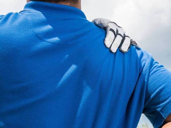 A man with a blue shirt holds a golf club in one hand and touches his right shoulder with his left hand while he looks out toward the golf course.