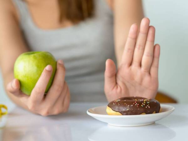 Woman using one hand to resist eating a doughnut while holding a healthy apple that she plans to eat.