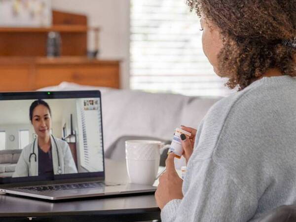 A woman sits at a table while she meets with her doctor virtually on a laptop computer and discusses medication.