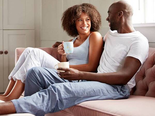 A young black man and woman smile as they sit relaxed on the couch with their feet up.