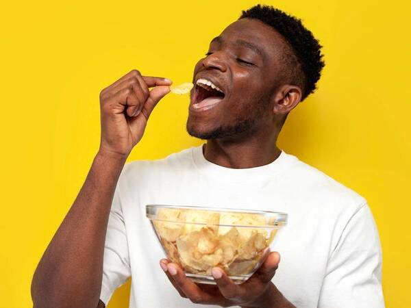 A young black man holds a bowl of chips and is about to put a chip in his mouth.