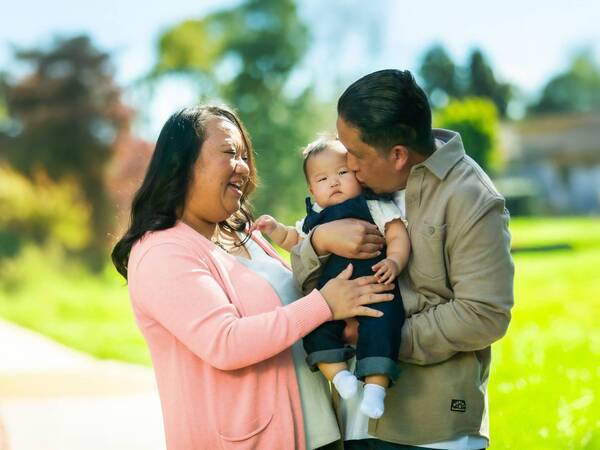 Mai Vue and husband Richard kiss their new baby girl Athena outdoors on a sunny day at a park.