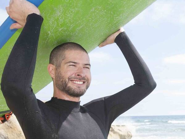Robert Elliott, DO, an internal medicine physician at Scripps Coastal Medical Center Hillcrest, pictured holding a surfboard at the ocean in San Diego.