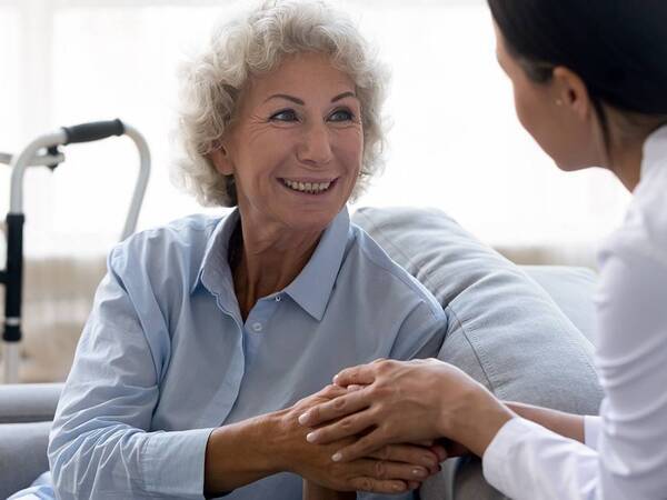 A senior woman on a sofa hold hands with a caregiver. A walker used for balance is in the background.