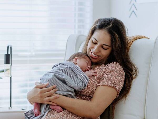 A mother smiling at her sleeping newborn sitting on a couch.