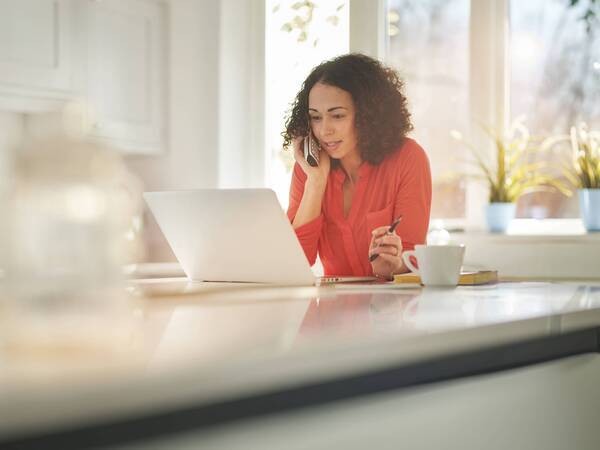 A young woman talks on the phone while looking at her laptop in her kitchen.