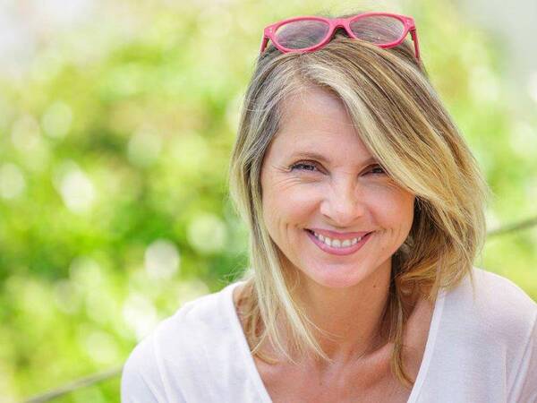 A casually dressed, smiling woman enjoys a quiet moment in a sunny outdoor setting.
