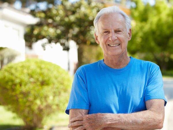 A casually dressed, smiling elderly gentleman relaxes while standing in his driveway on a sunny day.